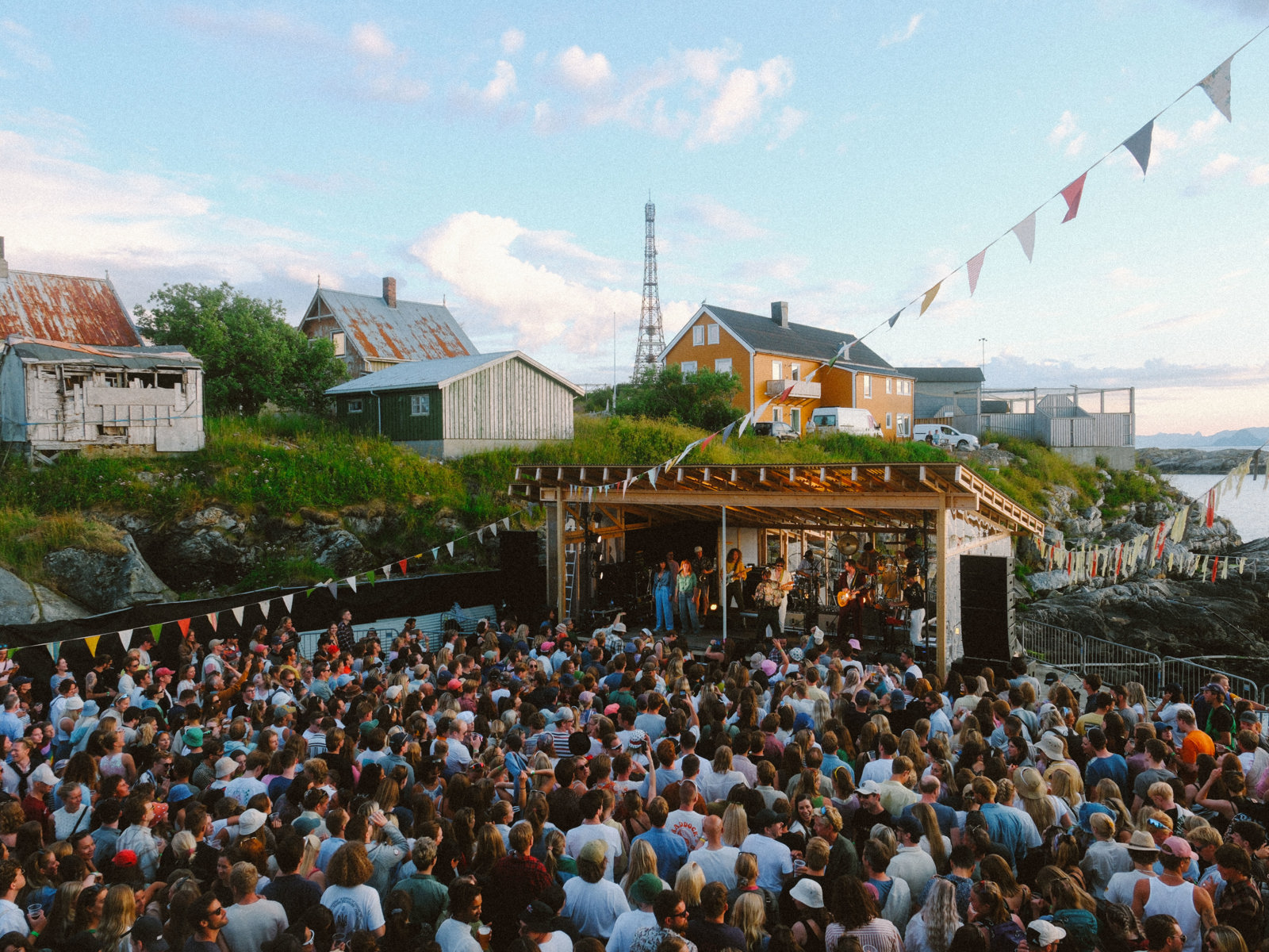 Foto av konsert utendørs i sommerkveldinga. Masse mennesker, vimpler, enebolig i bakgrunnen. 