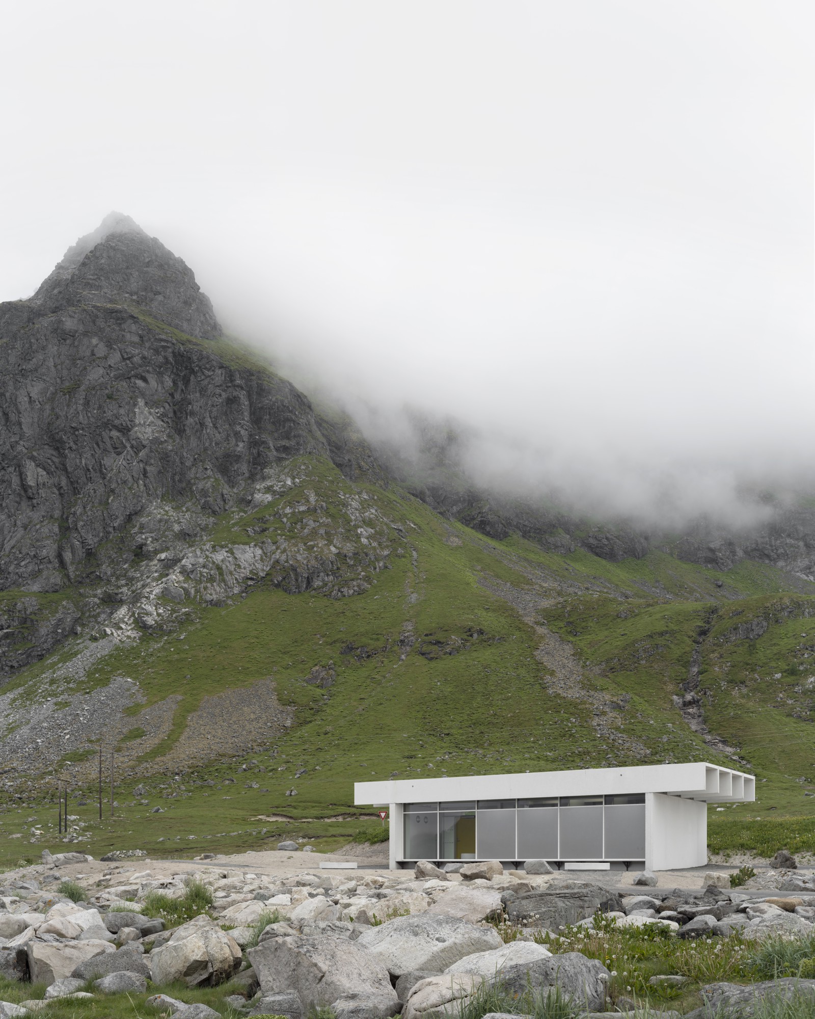 Foto av lite hvitt servicebygg midt i landskapet, fjell og skyer i bakgrunnen.