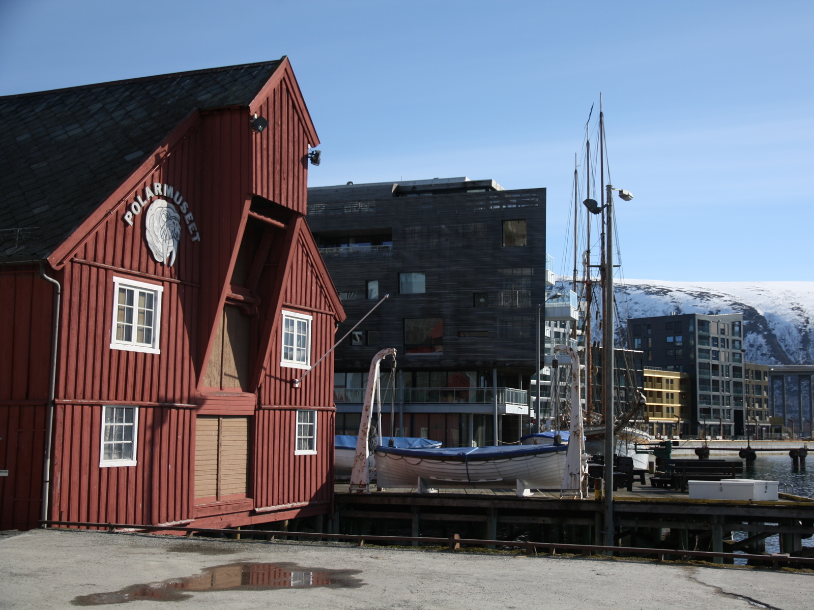 Foto av bryggekant, tradisjonelt rødt bygg i forgrunnen, moderne leilighetsblokker i bakgrunnen, fjell bak der igjen. 