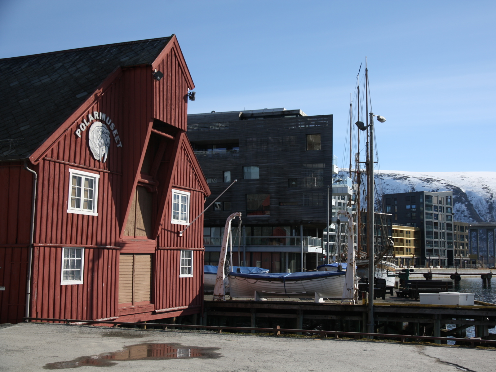 Foto av bryggekant, tradisjonelt rødt bygg i forgrunnen, moderne leilighetsblokker i bakgrunnen, fjell bak der igjen.