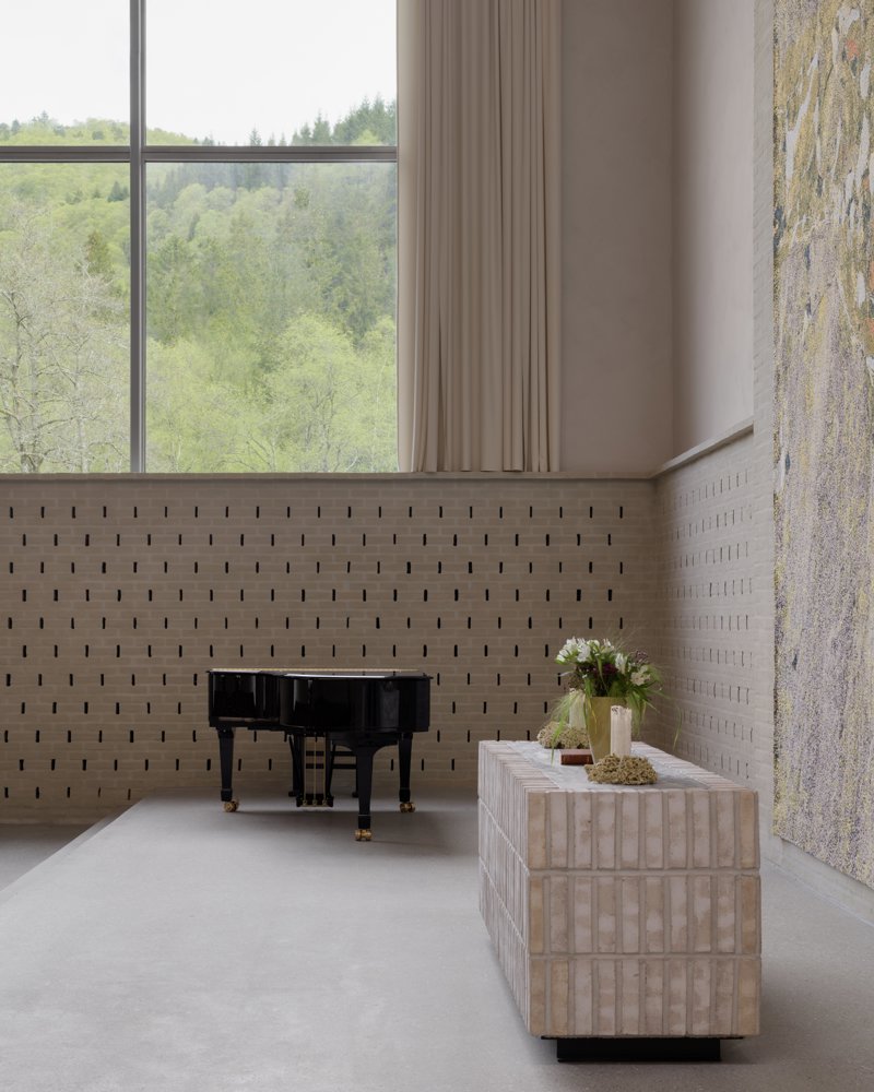 Foto av alter i lys teglstein, flygel og et stort vindu ut mot skogen i bakgrunnen.