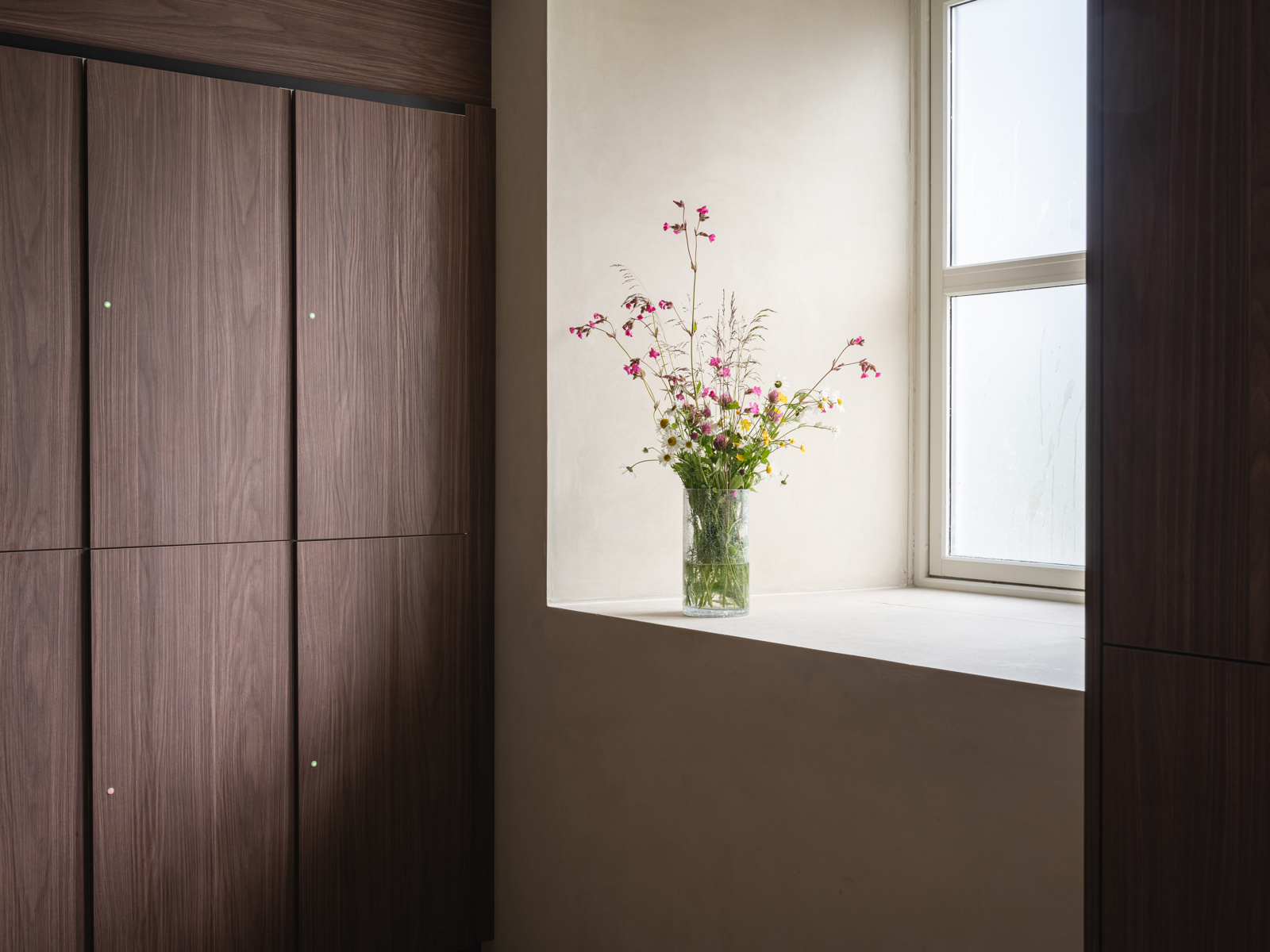 Interiør foto av garderobe, skap i mørkt treverk, vindu med en vase blomster i karmen.