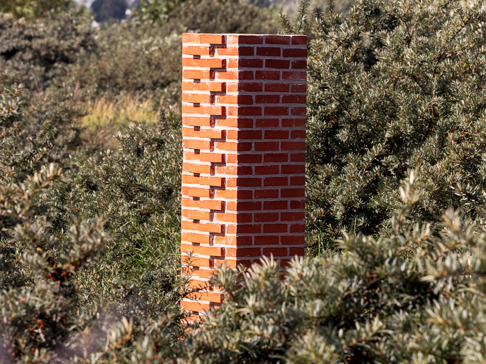 En murt søyle i rød teglstein stående i lyng ute i naturen. Foto.