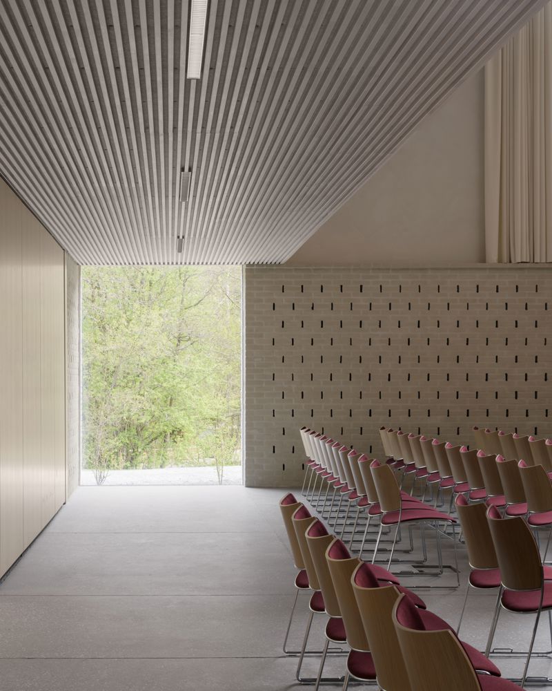 Interiør foto av bakerst i et lyst kirkerom, rader med stoler og et stort vindu ut til skogen.