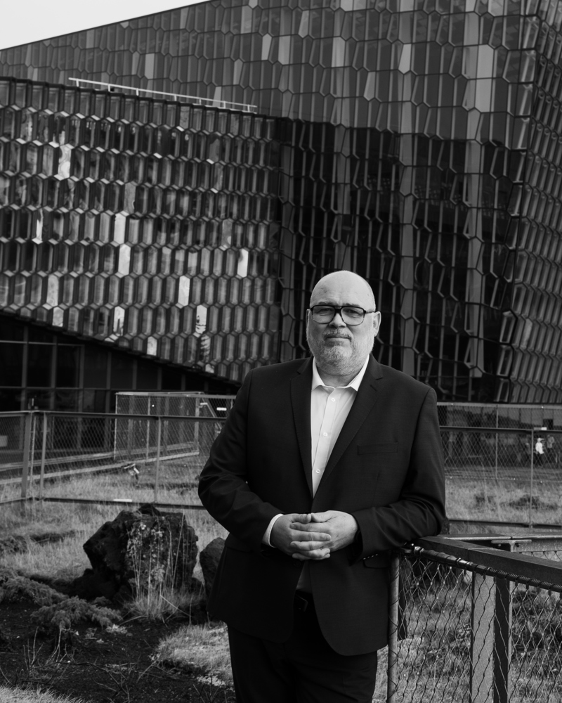 Portrett foto av Logi Már Einarsson med et stort bygg i bakgrunnen, svart hvitt.