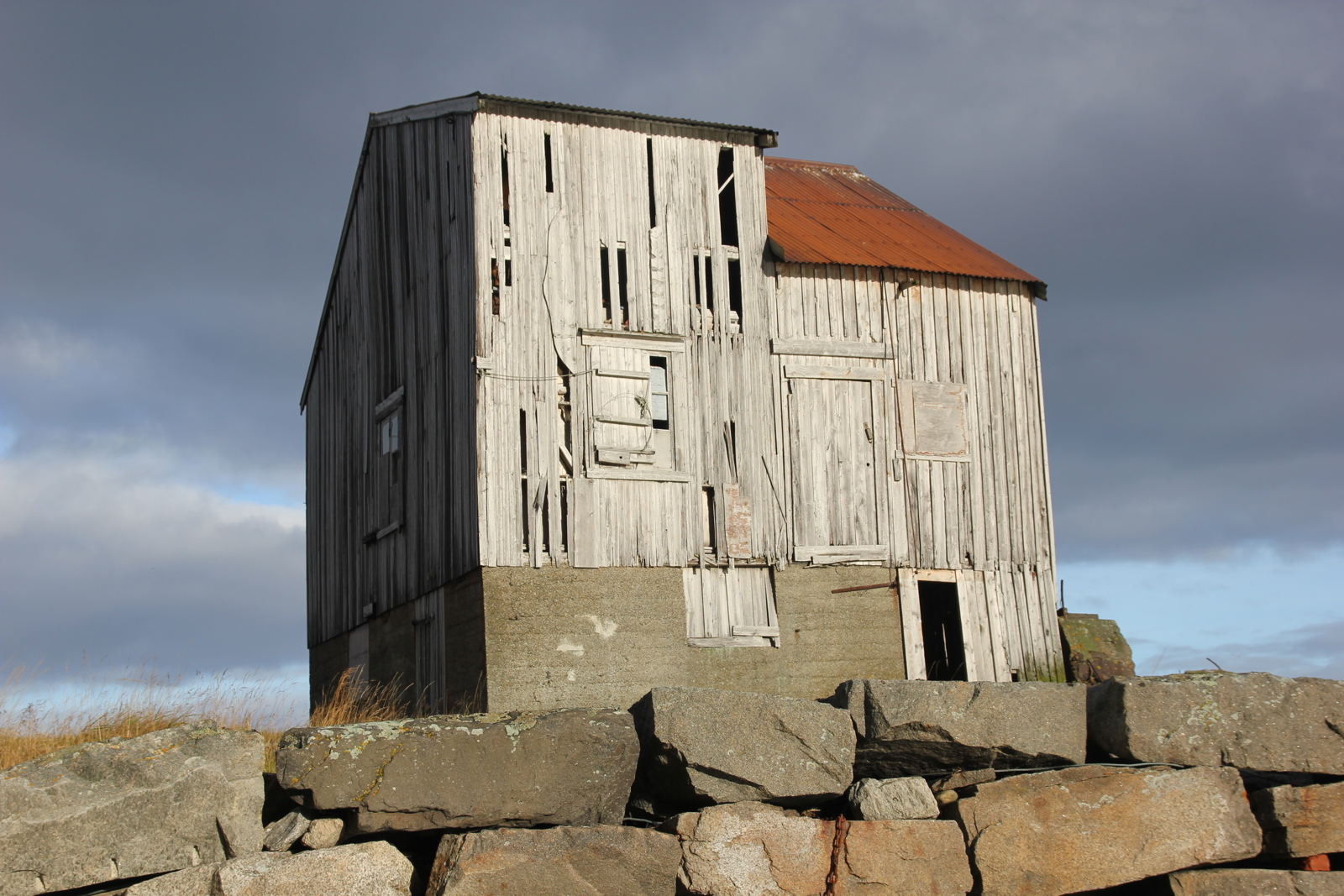 Foto av nedslitt grånet hus, steinmur i forkant. 