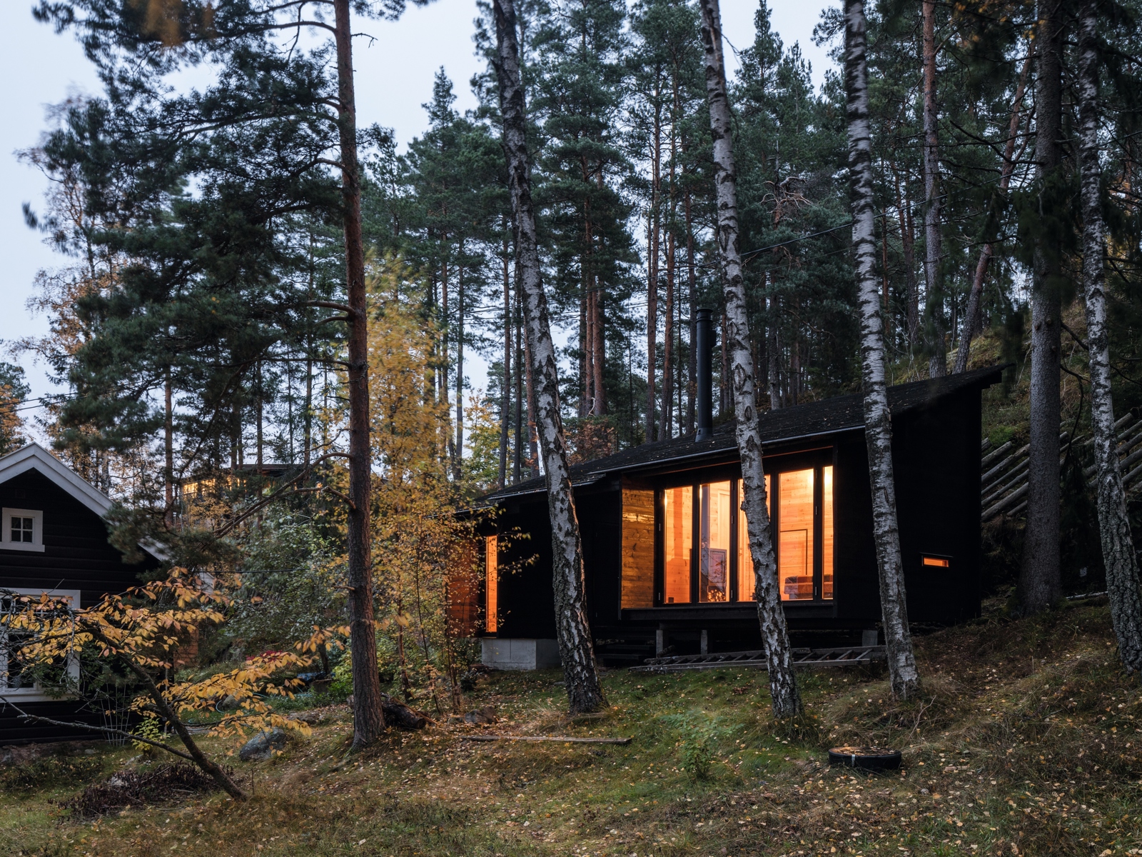 Eldre hytte ved siden av nyere hytte i laft som lyser opp i skumringen. Skog rundt. Eksteriør Foto.