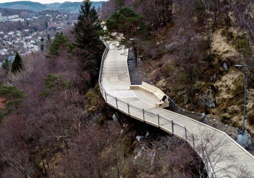 Oversikts foto/ dronefoto av gangsti i treverk i åsside, med by i bakgrunnen.