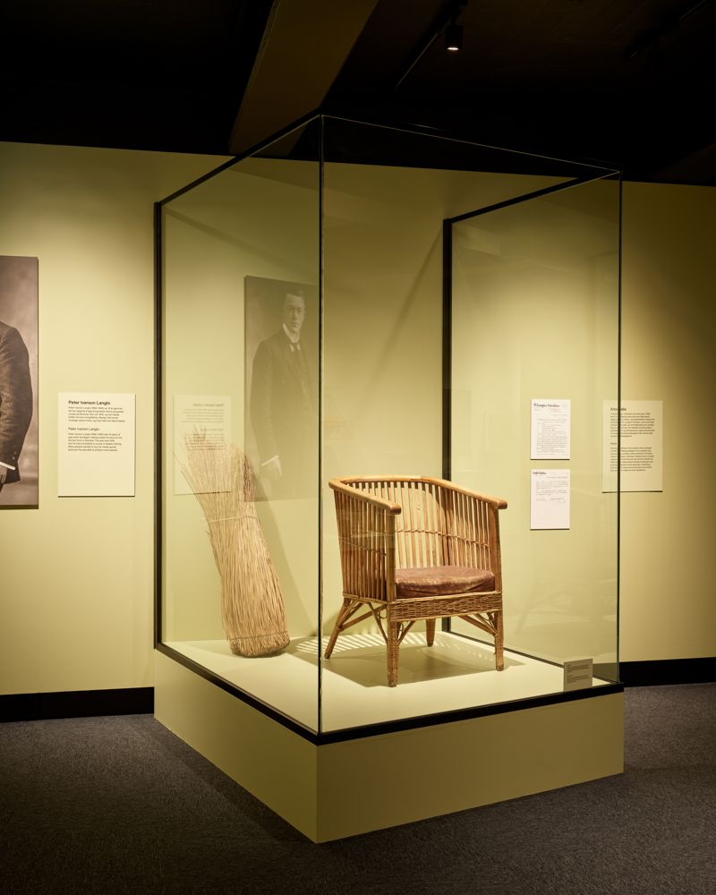 En stol i en glassmonter, gul vegg bak. Utstilling. Foto.