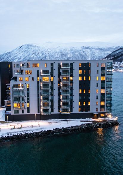 Foto av boligblokk på tuppen av havn, vann rundt, snøkledde fjell i bakgrunnen, vinter lys. 