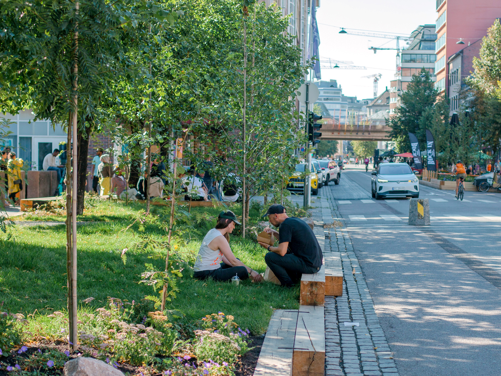 Foto av en gate med grønn plen og trær på en side og bilvei på andre siden, et par sitter og spiser på en benk og gresset.