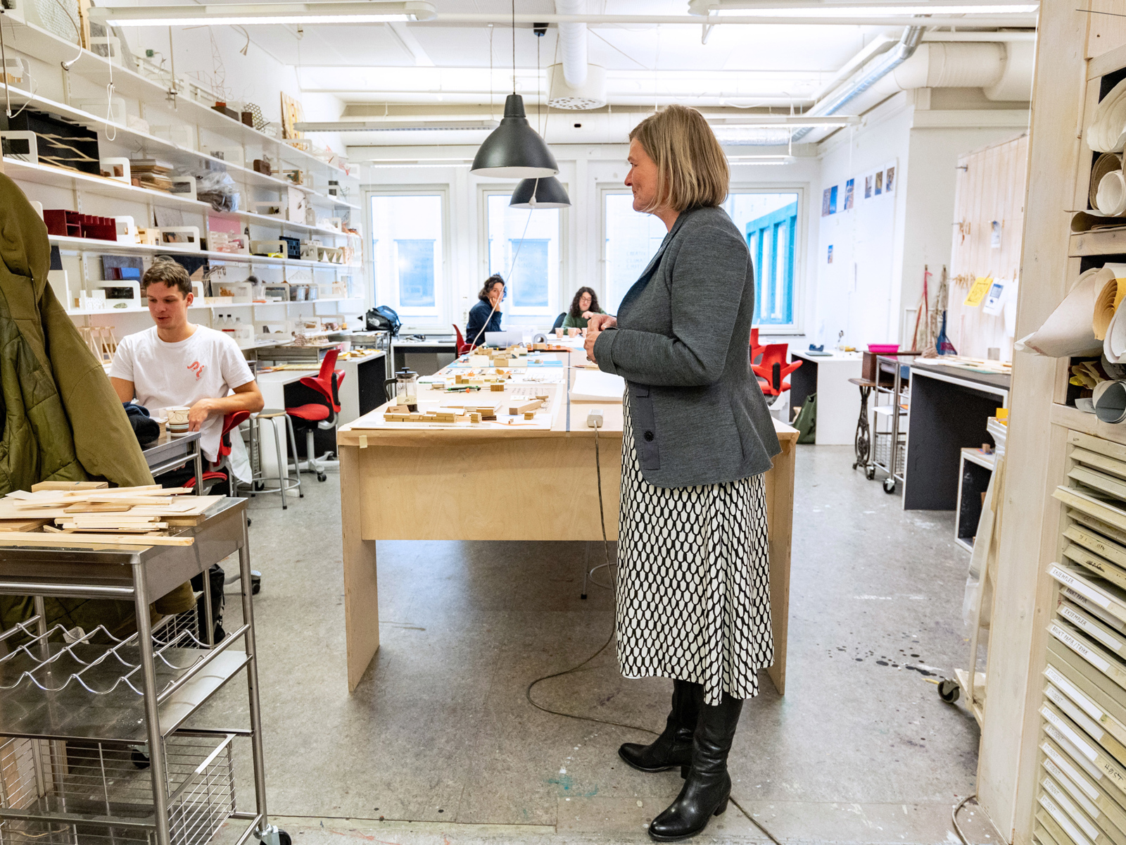 Marianne Skjulhaug står i et rom med elever og arbeidsbord fullt med ting. Foto.