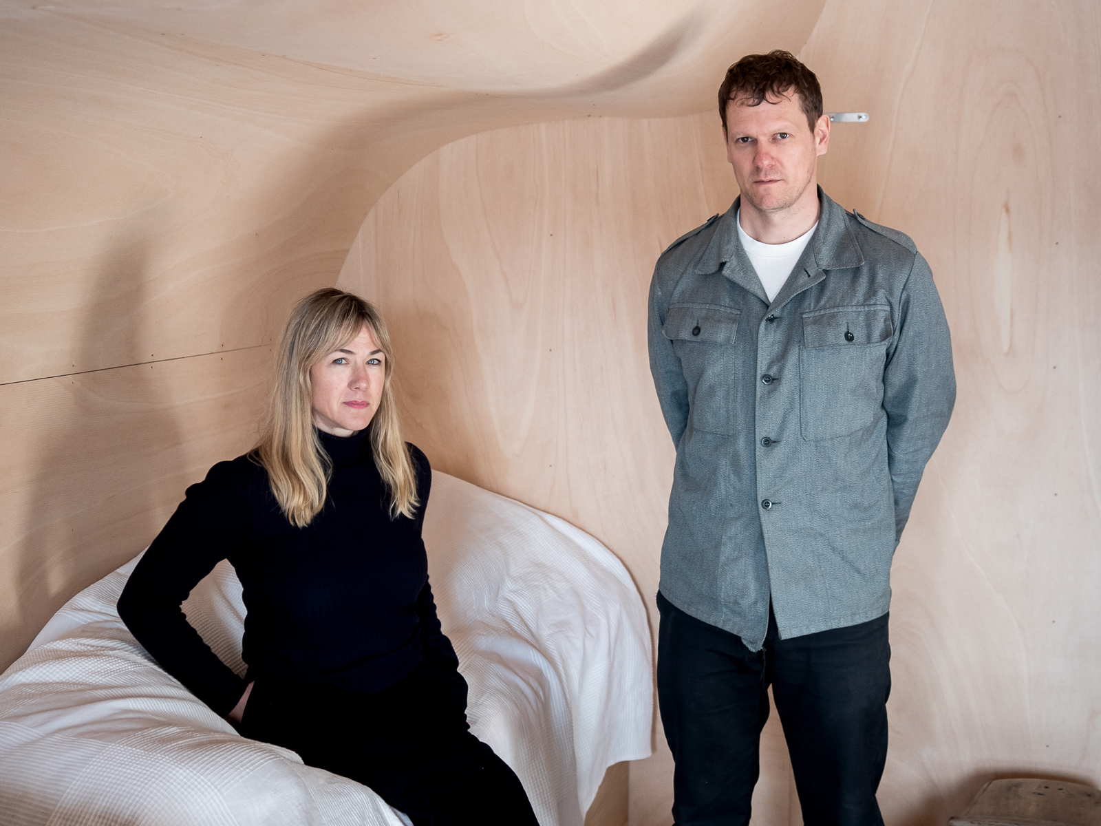 Marina Bauer sitter på ett hvitt mykt objekt og Espen Folgerø står. Rommet er av tre og taket bølger og blir en del av veggen. Foto.